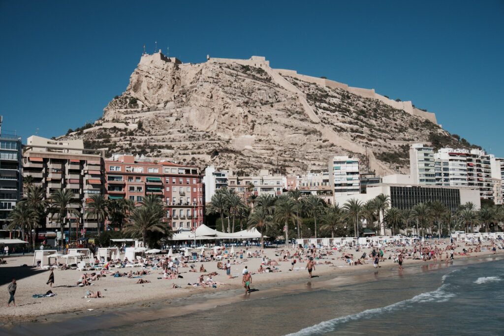 Beliebter Stadtstrand von Alicante mit Blick auf die bunte Strandpromenade und die Burg Castillo de Santa Bárbara, Costa Blanca, Spanien.