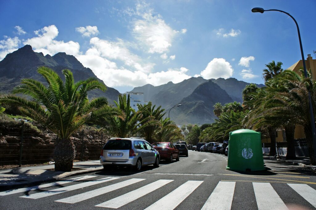 Straße mit Palmen und geparkten Autos vor der Bergkulisse im Ort Los Gigantes, Teneriffa, Kanarische Inseln.