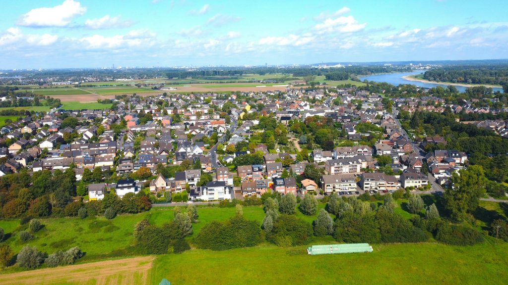 Luftaufnahme von Zons am Rhein mit Wohnhäusern, Grünflächen und dem Rhein im Hintergrund.