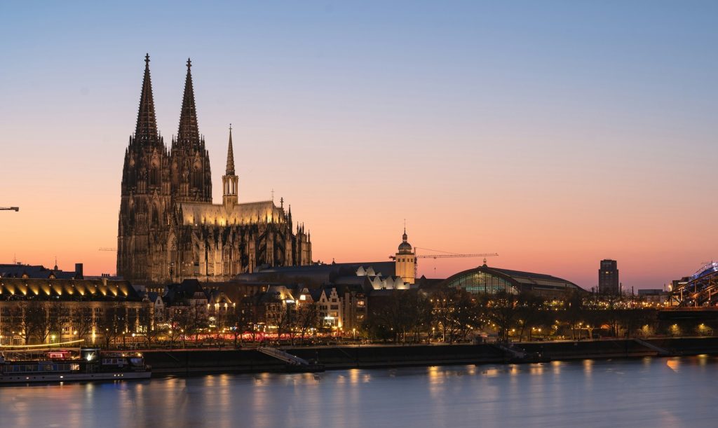 Kölner Dom in der Abenddämmerung, beleuchtet vor orangefarbenem Himmel, mit Rheinspiegelung im Vordergrund.