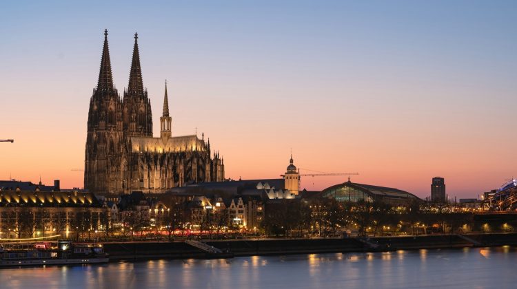 Kölner Dom in der Abenddämmerung, beleuchtet vor orangefarbenem Himmel, mit Rheinspiegelung im Vordergrund.