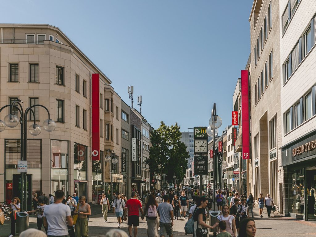 Belebte Fußgängerzone in Köln mit Geschäften und Passanten an einem sonnigen Tag.