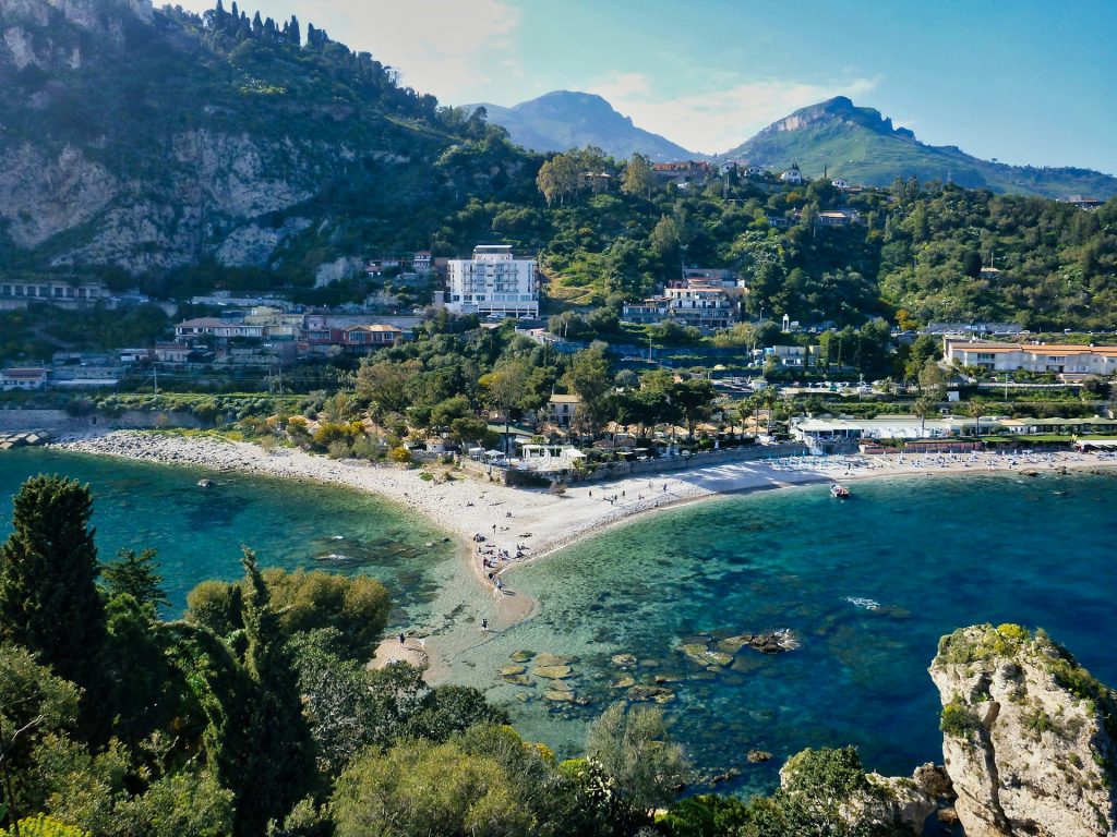 Luftaufnahme der Bucht von Taormina mit türkisblauem Meer, Sandstrand und Bergen im Hintergrund.