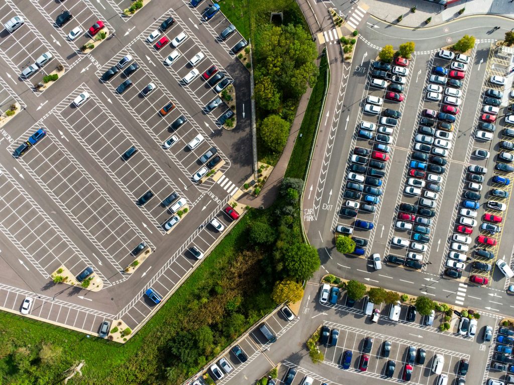 Drohnenaufnahme zweier großer Außenparkplätze mit farbigen Fahrzeugen und Grünstreifen dazwischen.