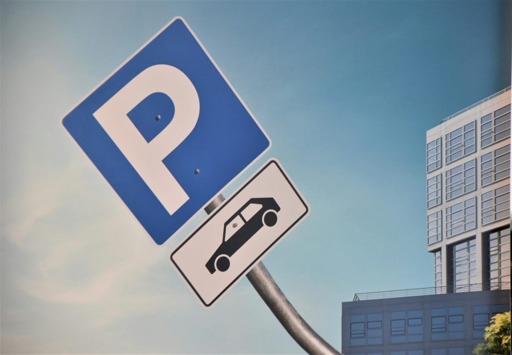 Blaues Parkschild mit Auto-Piktogramm vor modernem Gebäude und blauem Himmel.