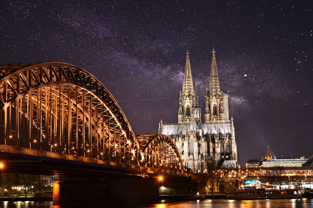Hohenzollernbrücke und beleuchteter Kölner Dom bei Nacht unter einem Sternenhimmel.