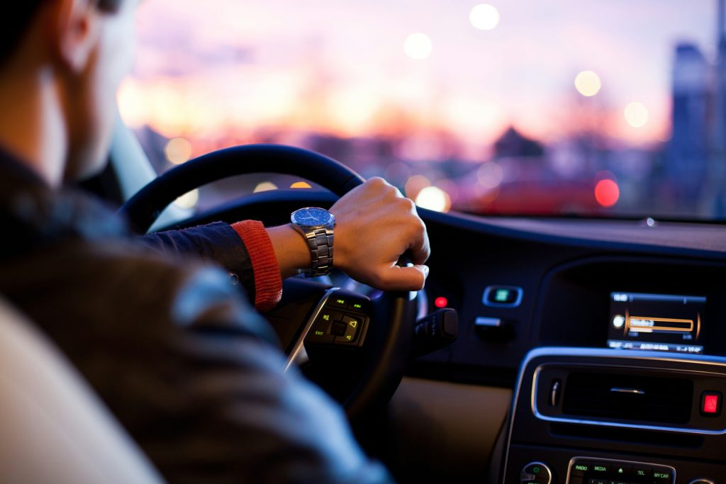 Mann am Steuer eines Autos bei Nacht, Armbanduhr am Handgelenk, verschwommene Stadtlichter im Hintergrund.