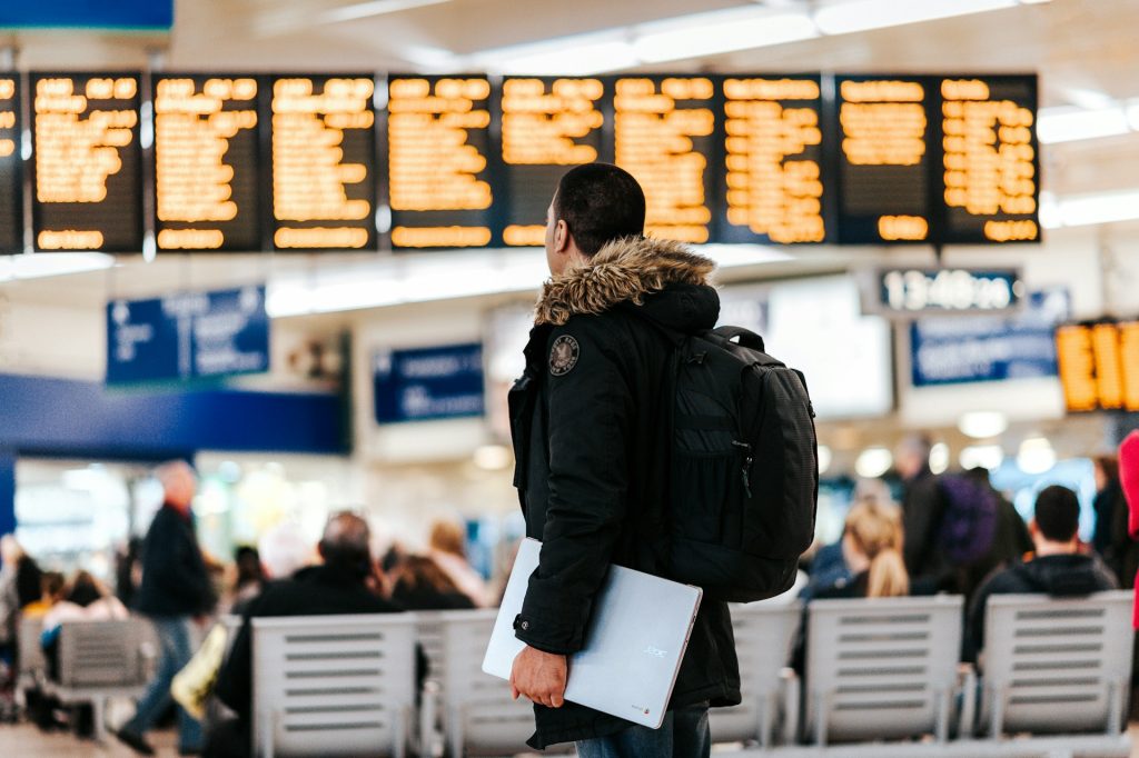 Mann mit Rucksack und Laptop betrachtet eine beleuchtete Abflugtafel in einem belebten Flughafenterminal.
