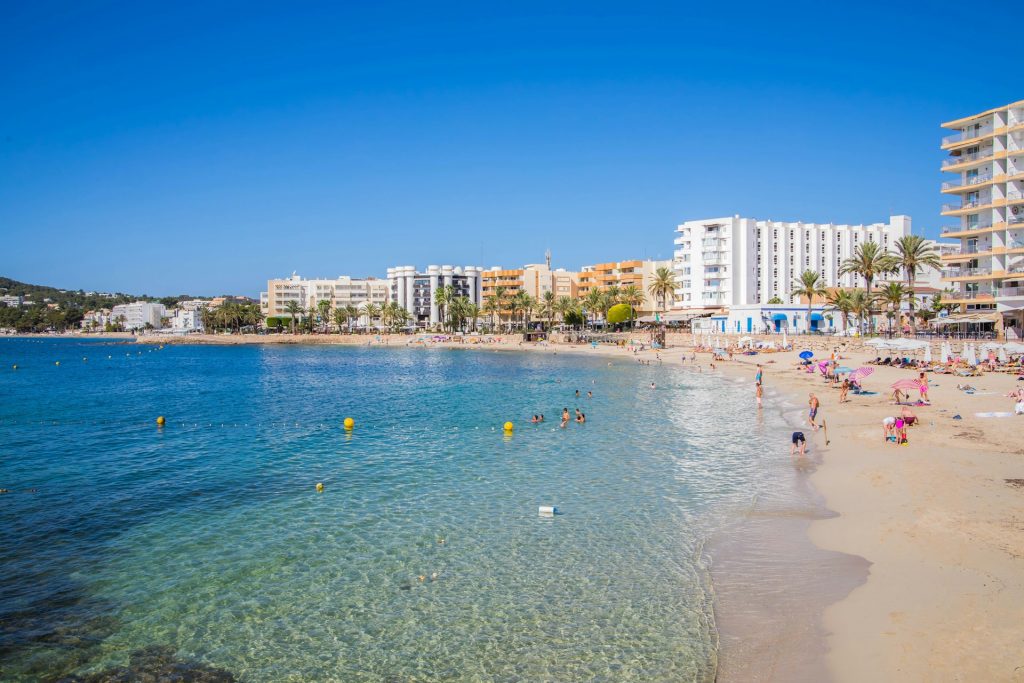 Belebter Sandstrand in Ibiza mit türkisblauem Wasser, Palmen und weißen Hotels im Hintergrund.