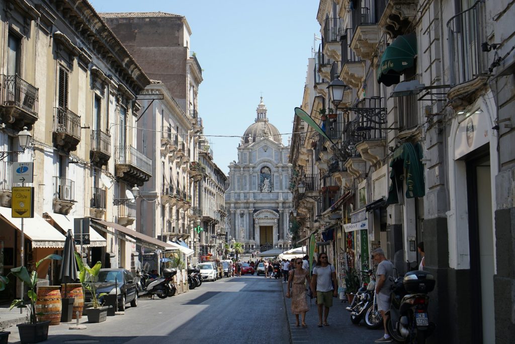 Belebte Straße in Catania mit Passanten, Cafés und barockem Dom im Hintergrund.