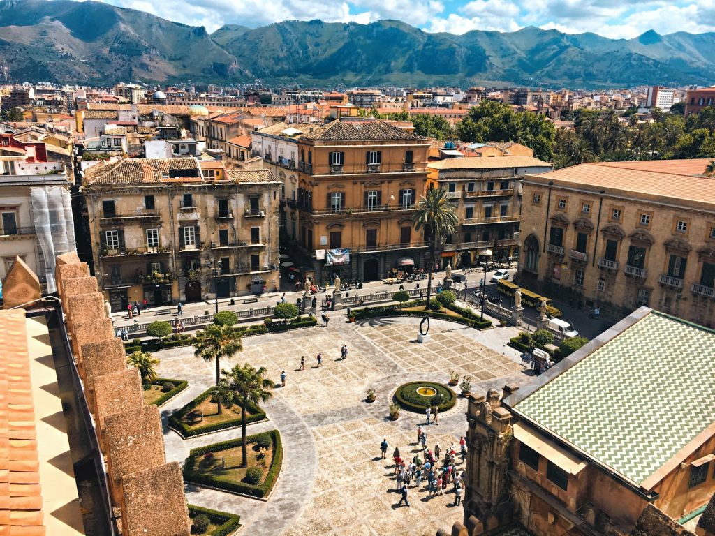 Luftaufnahme eines belebten Platzes in Palermo mit historischen Gebäuden, Palmen und Bergen im Hintergrund.
