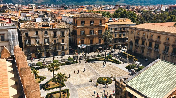 Luftaufnahme eines belebten Platzes in Palermo mit historischen Gebäuden, Palmen und Bergen im Hintergrund.