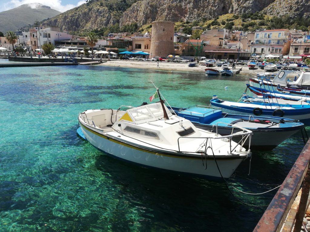 Fischerboote im türkisblauen Hafen von Mondello mit Bergen und historischem Turm im Hintergrund.