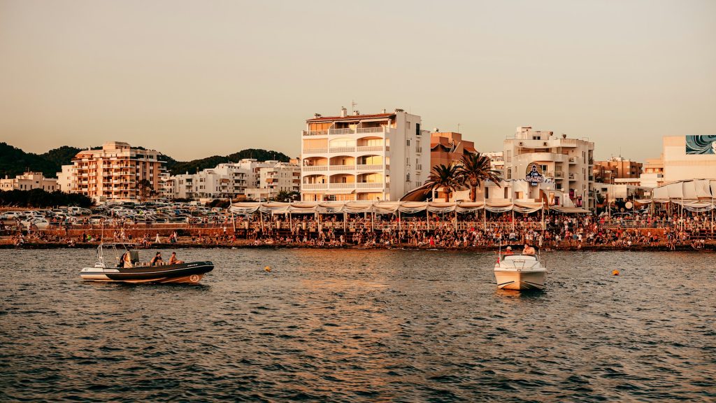 Boote auf dem Meer vor der belebten Strandpromenade von Ibiza bei Sonnenuntergang.