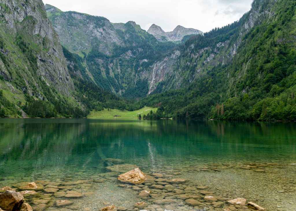 Kristallklarer Königssee mit smaragdgrünem Wasser, umgeben von steilen Felswänden und einem Wasserfall im Hintergrund.