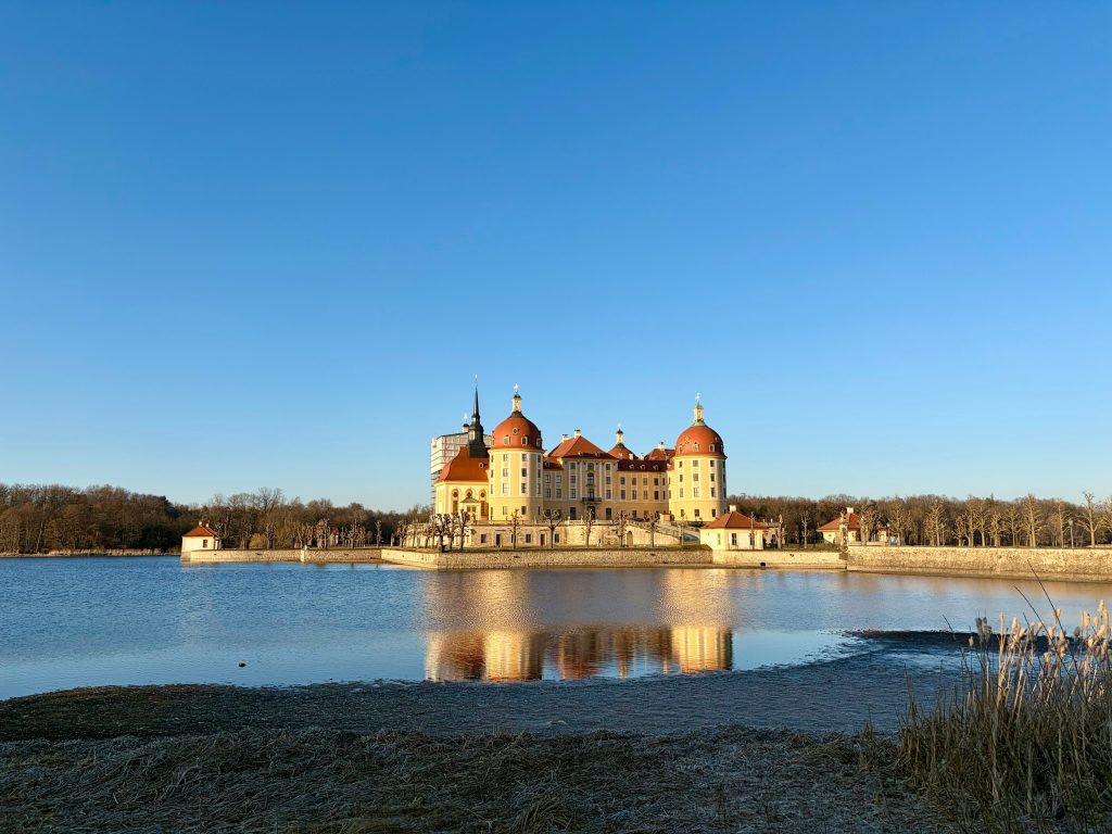 Barockschloss Moritzburg auf einer Insel mit Spiegelung im ruhigen Schlossteich unter blauem Himmel.