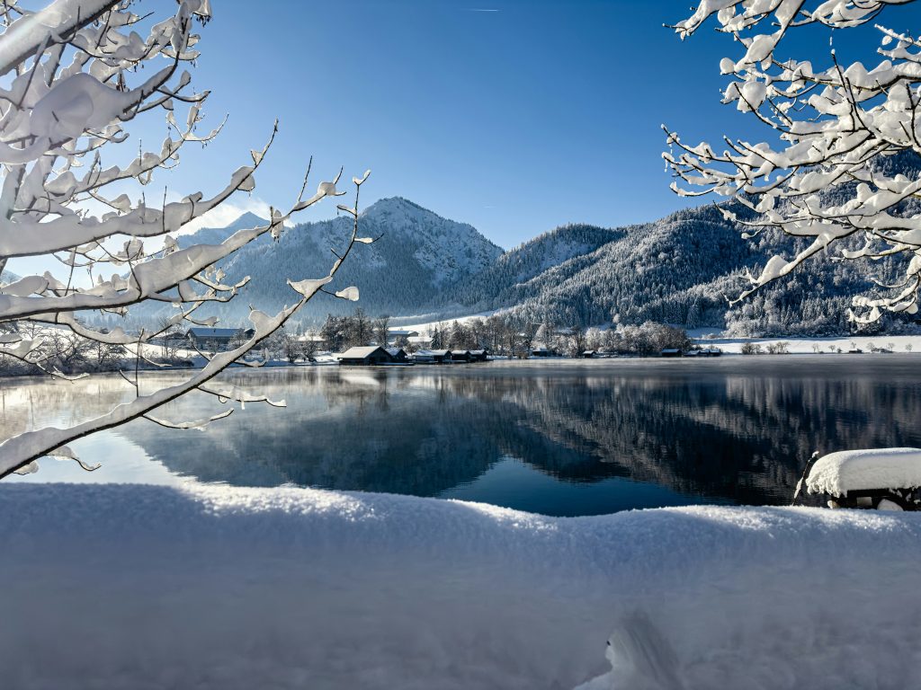 Winterlicher Schliersee mit schneebedeckten Bäumen, Bergen und ruhiger Wasseroberfläche unter blauem Himmel.