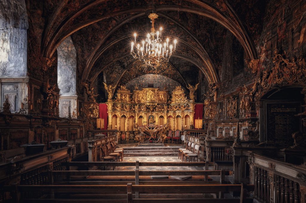 Prunkvoller Innenraum einer historischen Kapelle mit goldenem Altar, Holzbänken und einem Kronleuchter.