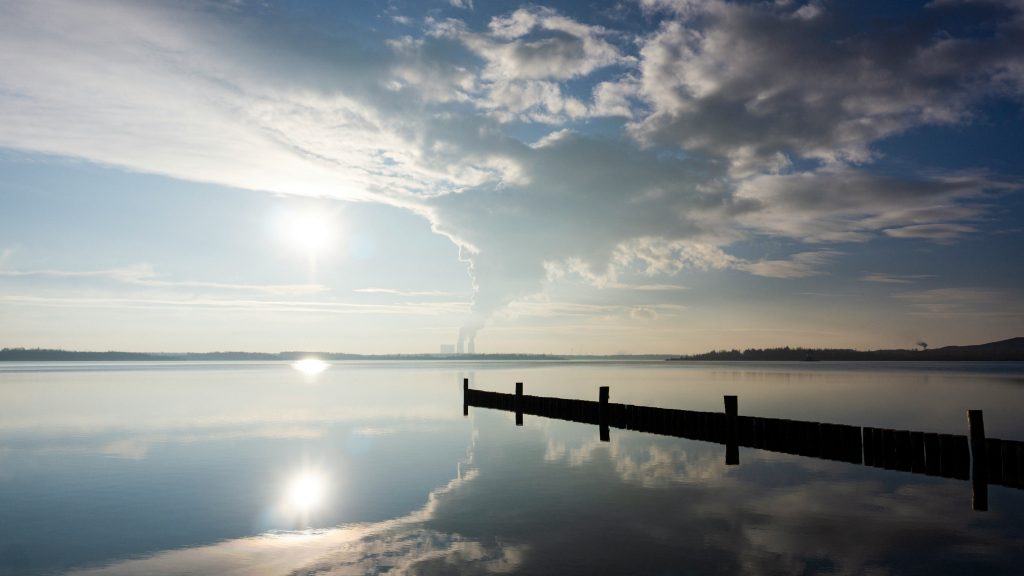 Ruhige Wasseroberfläche des Störmthaler Sees mit einem Holzsteg und Sonnenspiegelung unter bewölktem Himmel.