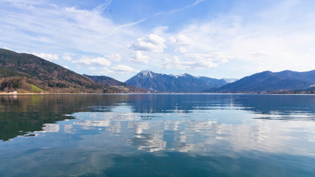 Gelbes Ruderboot an einem Holzsteg am Chiemsee mit Segelbooten und den Alpen im Hintergrund.