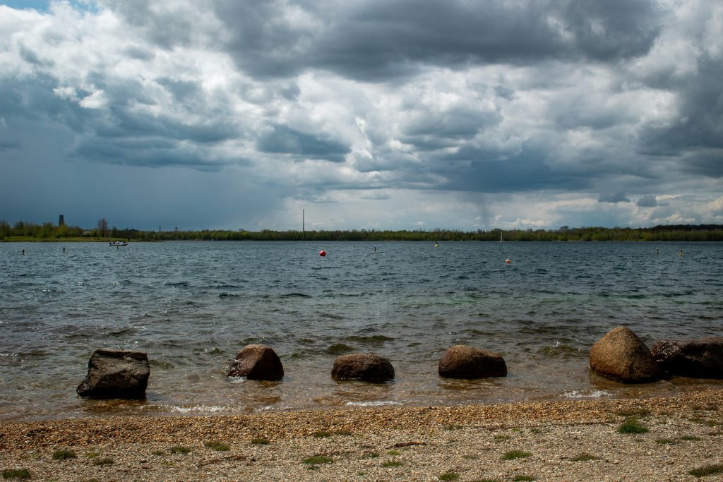 Steiniger Uferstreifen am Cospudener See mit bewölktem Himmel und Bojen auf dem Wasser.