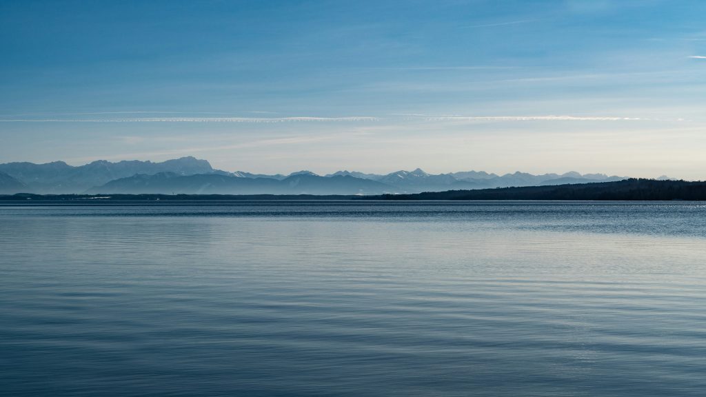 Ruhige Wasseroberfläche des Starnberger Sees mit schneebedeckten Alpen im Hintergrund.