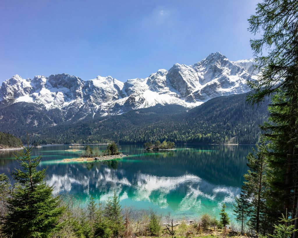 Türkisfarbener Eibsee mit schneebedeckten Zugspitze-Gipfeln und Nadelwäldern unter blauem Himmel.
