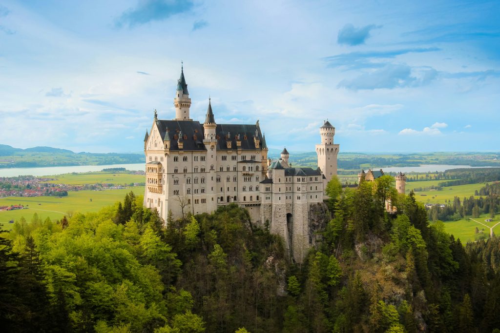 Schloss Neuschwanstein auf einem bewaldeten Felsen bei Füssen mit grüner Landschaft und See im Hintergrund.